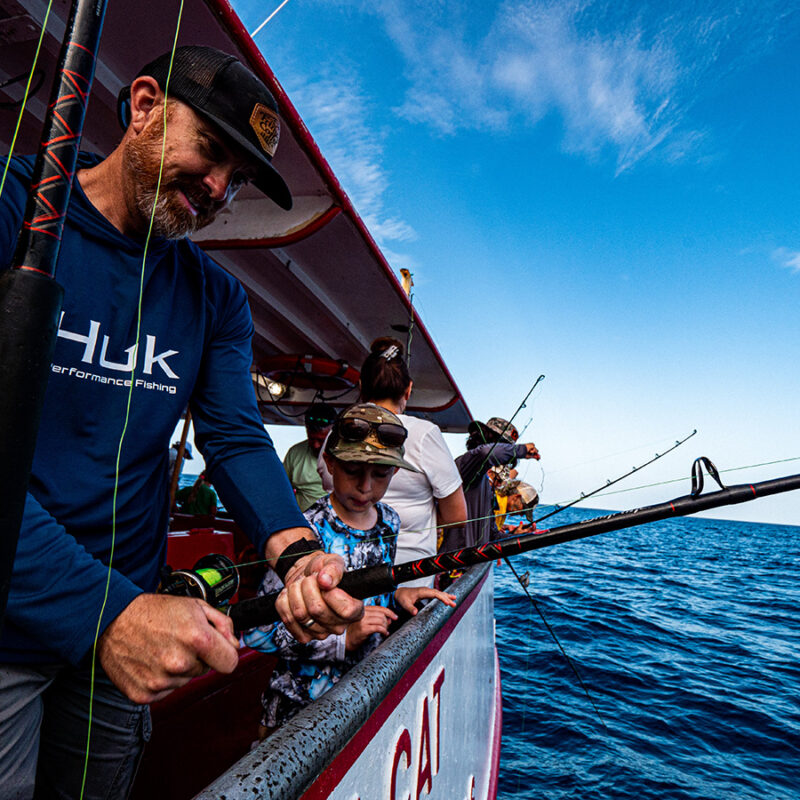 Family fishing trip with an adult assisting a young child with their fishing rod on a charter boat, against a backdrop of open sea under a blue sky