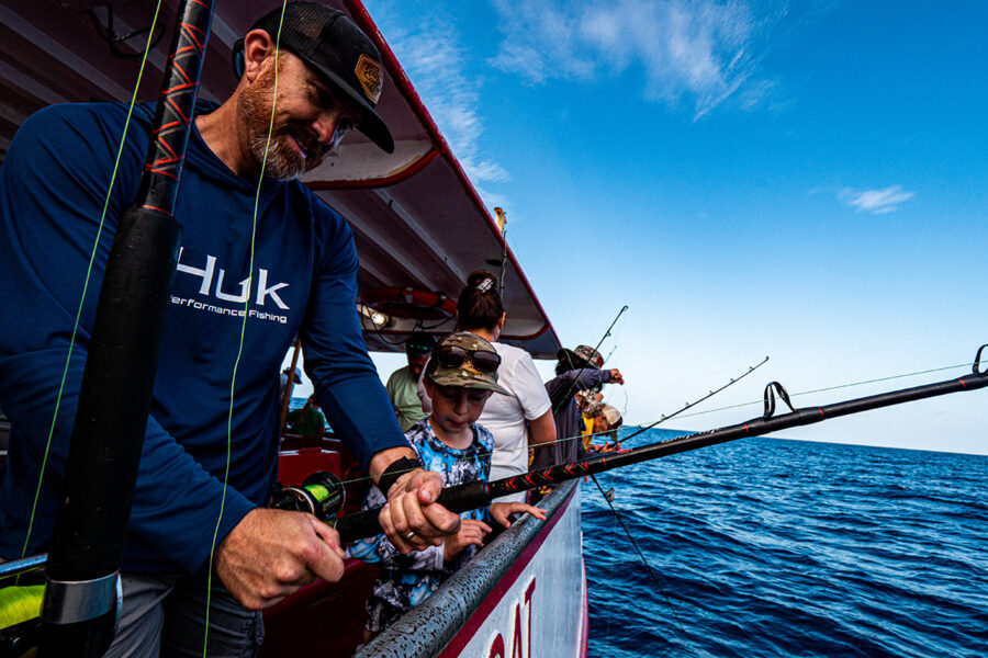 Family fishing trip with an adult assisting a young child with their fishing rod on a charter boat, against a backdrop of open sea under a blue sky