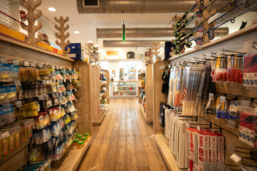 Aisle view inside a fishing supply store with wooden shelves stocked with various fishing lures, tackle, and accessories, featuring wooden floor and maritime-themed decorations.