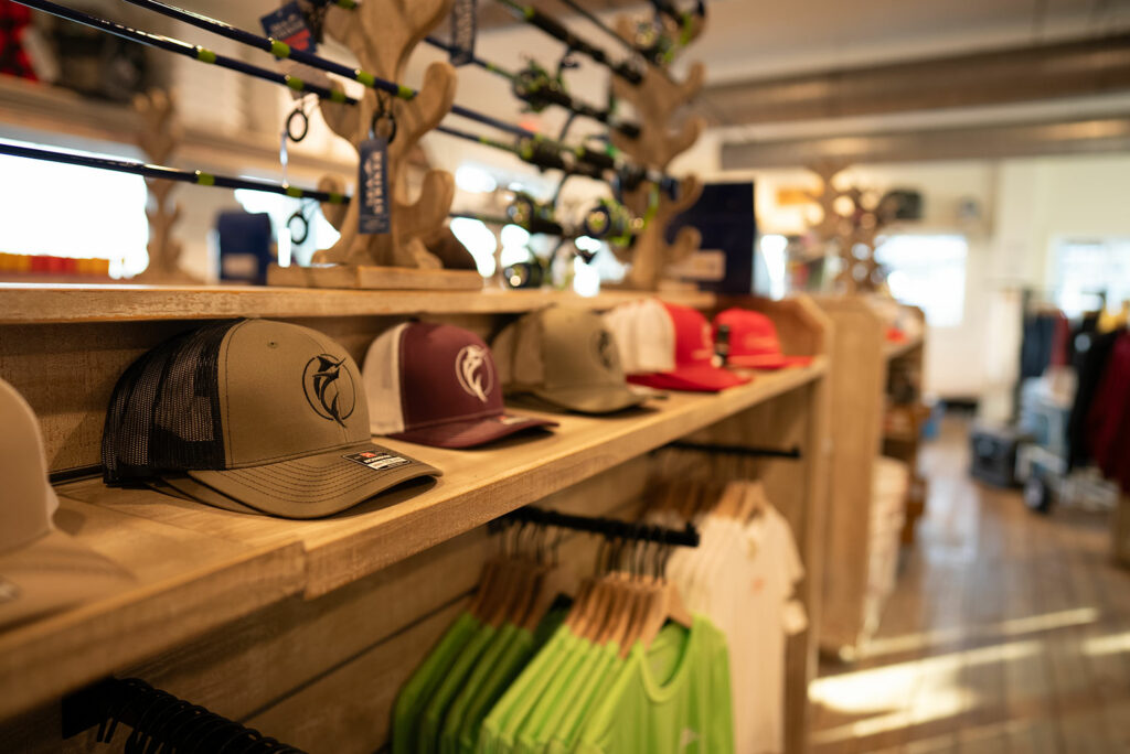 Interior of Port Aransas Fisherman's Wharf marina with a display of various caps on a wooden shelf, fishing rods hanging in the background, and other maritime apparel visible.